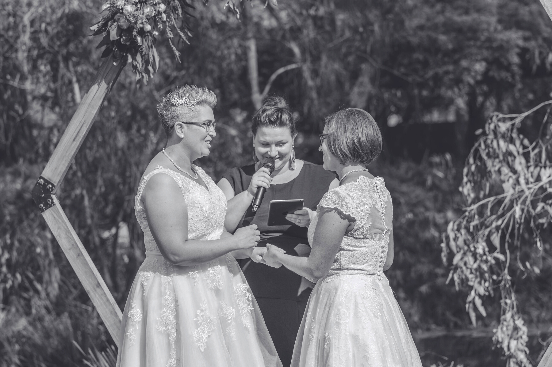 Two brides holding hands and smiling at each other. A Marriage Celebrant stands between them smiling looking down at her device talking into her microphone.